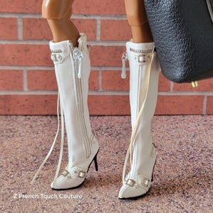 Puede incluir: Botas blancas hasta la rodilla con cremalleras y hebillas. Las botas tienen punta y tacones de aguja. Un bolso negro es visible en el fondo. Las botas est&aacute;n en una mu&ntilde;eca.
