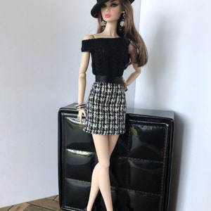 May include: A fashion doll wearing a black off-the-shoulder sweater, a black and white checkered skirt, a black hat, and black shoes. The doll is standing next to a black box with a quilted pattern.