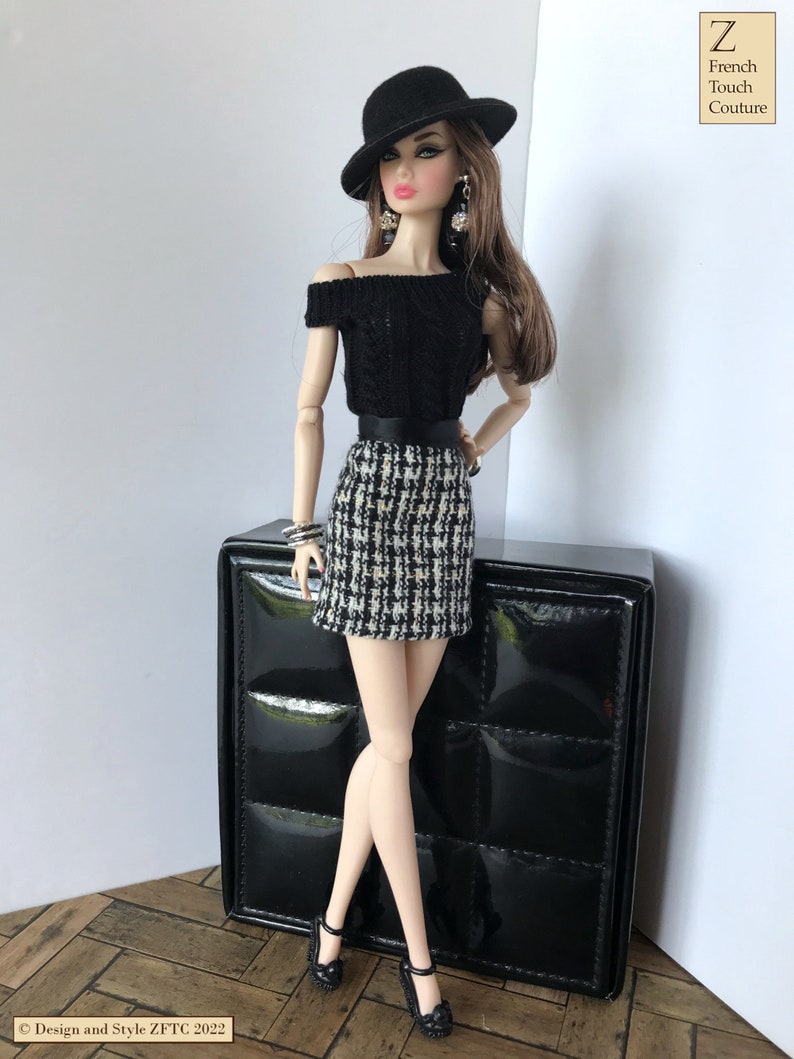 May include: A fashion doll wearing a black off-the-shoulder sweater, a black and white checkered skirt, a black hat, and black shoes. The doll is standing next to a black box with a quilted pattern.