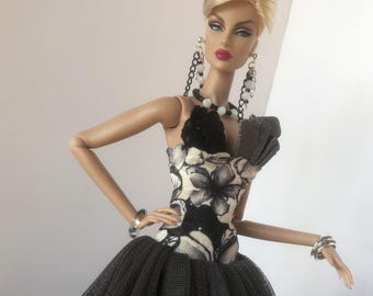 May include: A fashion doll wearing a black and white floral print dress with a black tulle skirt. The doll has blonde hair styled in a bouffant and is wearing silver earrings and a silver bracelet.