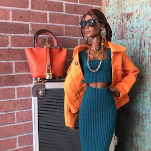 May include: A doll wearing a teal ribbed knit crop top and pencil skirt, paired with an orange jacket and sunglasses. A small orange handbag with a gold lock sits on a suitcase. The doll is accessorized with gold jewelry and earrings.