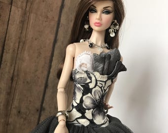 May include: A fashion doll with long brown hair, wearing a black and white floral print dress with a layered tulle skirt. The doll is accessorized with silver jewellery, including earrings, a necklace, and a bracelet. The doll has a fair complexion and is posed against a wooden background.