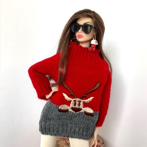 May include: A fashion doll with long brown hair wearing black sunglasses, a red and gray knit sweater featuring an embroidered reindeer graphic, and a dangling earring shaped like a red Christmas stocking with white trim.