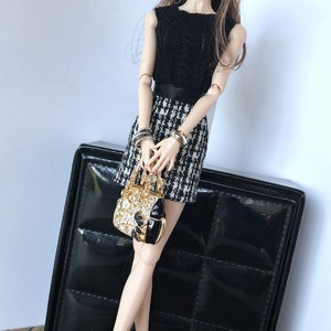 May include: A fashion doll wearing a black and white houndstooth skirt, a black sleeveless top, a black hat, and gold jewelry. The doll is holding a gold handbag with rhinestones. The doll is standing next to a black box with a quilted pattern.