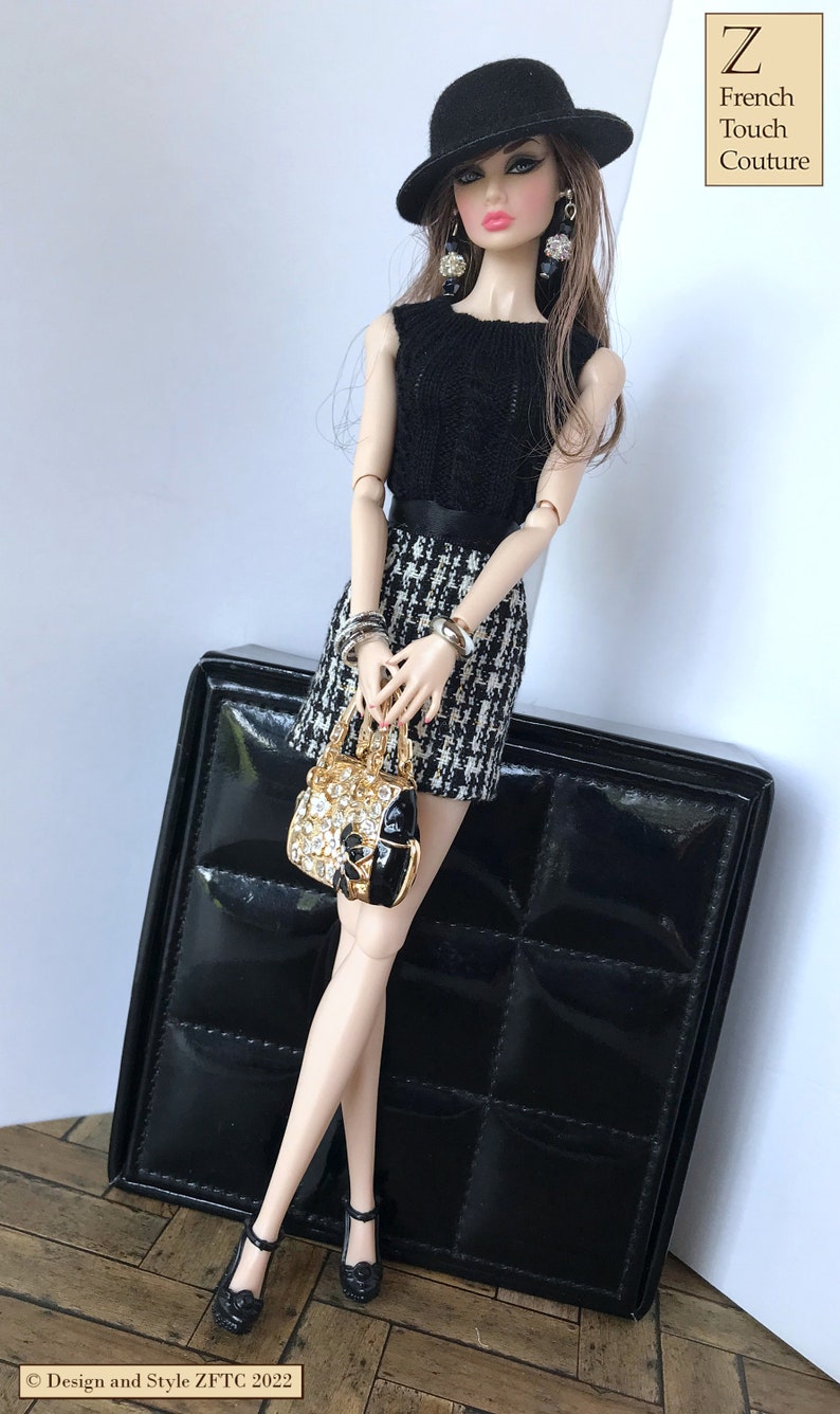 May include: A fashion doll wearing a black and white houndstooth skirt, a black sleeveless top, a black hat, and gold jewelry. The doll is holding a gold handbag with rhinestones. The doll is standing next to a black box with a quilted pattern.