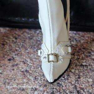 Puede incluir: Primer plano de una bota blanca con punta y tac&oacute;n peque&ntilde;o. La bota presenta hebillas plateadas decorativas y un dise&ntilde;o texturizado alrededor del tobillo. El texto "Z French Touch Couture" es visible en la parte inferior.