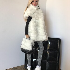 May include: A fashion doll wearing a white faux fur scarf, black top, grey and white patterned tights, and white boots. The doll is holding a white faux fur handbag and is standing next to a black quilted suitcase.