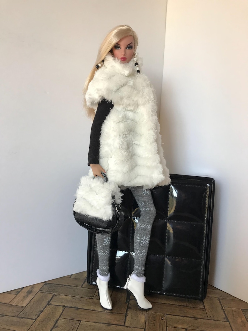May include: A fashion doll wearing a white faux fur scarf, black top, grey and white patterned tights, and white boots. The doll is holding a white faux fur handbag and is standing next to a black quilted suitcase.