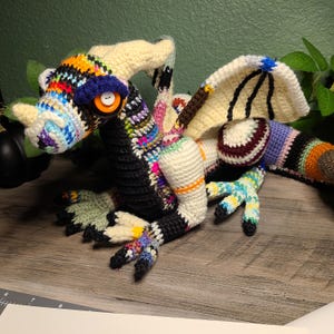 May include: A multi-colored crochet dragon plush toy featuring a black chest, cream-colored horns and wings, and a large orange button eye with a white center. The body is constructed from various yarn colors including blue, green, and purple.