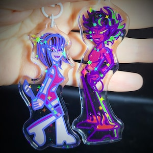May include: Two colorful character keychains with a holographic finish. The keychain on the left is a female character with purple hair, a red and blue outfit, and a knife. The keychain on the right is a male character with red skin, green hair, and a purple outfit. Both characters have stars and glitter accents.