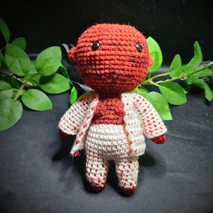 May include: A handmade crochet doll with a red head and body, wearing a white and red patterned jacket and pants. The doll has black eyes and small ears. The background is dark with green leaves.
