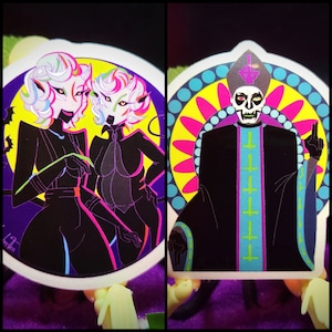 May include: Two black and white stickers with neon accents. The first sticker features two women in black outfits with neon green and pink accents. The second sticker features a figure in a black robe with a purple and green cross pattern. The figure has a white face with black eyes and a black crown.
