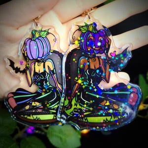 Op de afbeelding: Twee iriserende glitter-sleutelhangerbedels met een heks met een pompoenhoofd. De ene heks draagt een zwarte jurk met groene accenten en de andere draagt een zwarte jurk met paarse accenten. Beide heksen hebben een vleermuisvleugel en een hart op hun bedels.