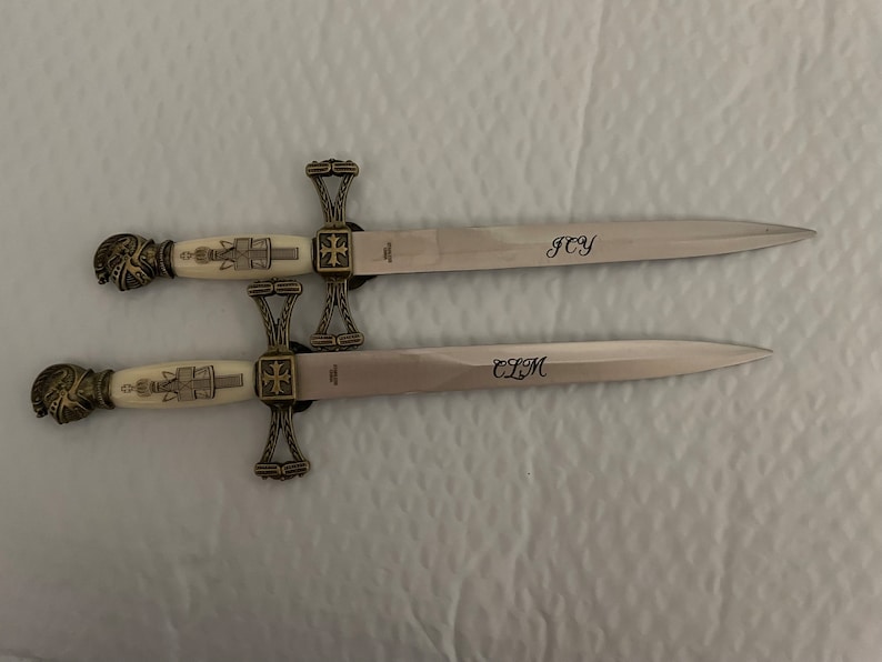 May include: Two decorative swords with gold-colored handles and cross guards. The blades are engraved with the initials "JCY" and "CLM".