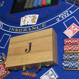 May include: A rectangular wooden poker chip case with a gold-colored handle and the letter J engraved on the lid. The case is surrounded by stacks of red, white, and blue poker chips in wooden racks, playing cards, and a blue blackjack table mat.