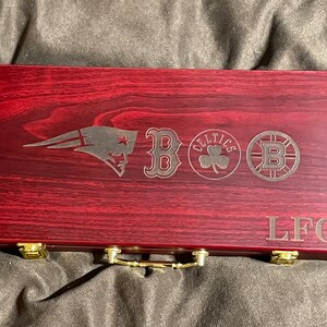 May include: A red wooden game box with a gold handle and brass accents. The box is engraved with the logos of the New England Patriots, Boston Red Sox, Boston Celtics, and Boston Bruins. The text "LFG" is also engraved on the box.