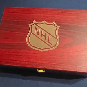 May include: A wooden box with a red mahogany finish. The box has a gold latch and a gold NHL logo engraved on the top.