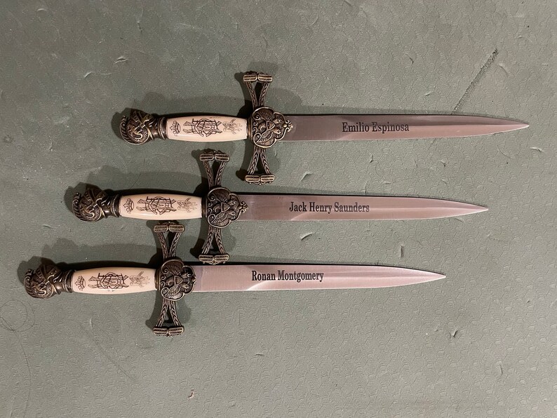 May include: Three ornate, silver-colored, ceremonial swords with ivory-colored handles. Each sword has a different name engraved on the blade: Emilio Espinosa, Jack Henry Saunders, and Ronan Montgomery.