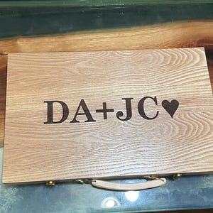 May include: A wooden box with a handle and a heart engraved on the front. The box is engraved with the text "DA+JC".