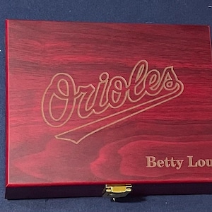 May include: A red wooden box with a gold latch. The box has an engraved Baltimore Orioles logo and the name "Betty Lou" on the front.