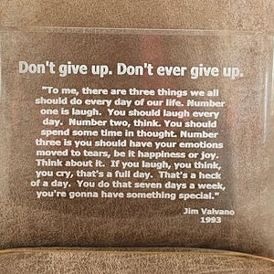 May include: A white acrylic plaque with a motivational quote by Jim Valvano, "Don't give up. Don't ever give up." The quote continues with a message about the importance of laughter, thought, and allowing yourself to feel emotions. The plaque is on a brown surface.