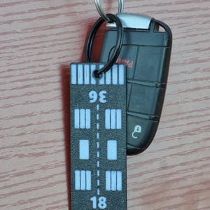 Könnte beinhalten: Schwarz-weißer Lineal-Schlüsselanhänger mit den Zahlen 18 und 36 darauf gedruckt. Der Schlüsselanhänger ist an einem schwarzen Schlüsselanhänger befestigt.