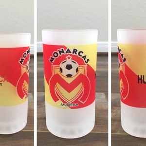 May include: A frosted glass beer stein with a red and yellow design featuring the logo of the soccer team "Monarcas Morelia". The logo includes a soccer ball and the team's name. The stein also has the text "Hasta la Muerte" and "Humberto" on it.