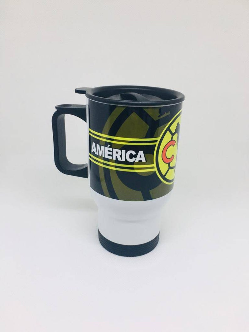 May include: A black and gold travel mug with a black lid and handle. The mug has a white stripe with the word "AM&Eacute;RICA" in yellow letters and a soccer ball logo with the letter "C" in red.