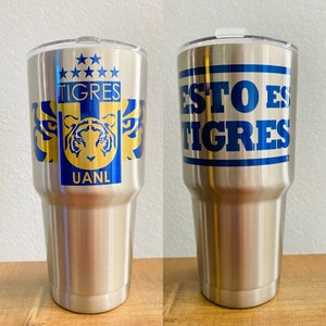 May include: Two stainless steel tumblers with blue and gold designs. One tumbler features the words "Tigres" and a tiger head logo. The other tumbler features the words "Esto es Tigres".