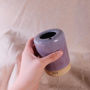 以下が含まれることがあります： 紫とベージュの色合いの円筒形のセラミック花瓶。花瓶は斑点のある仕上がりで、上部に広い開口部があります。底の部分はベージュで、ベースには数字の「333」が印刷されています。花瓶は手で持たれています。