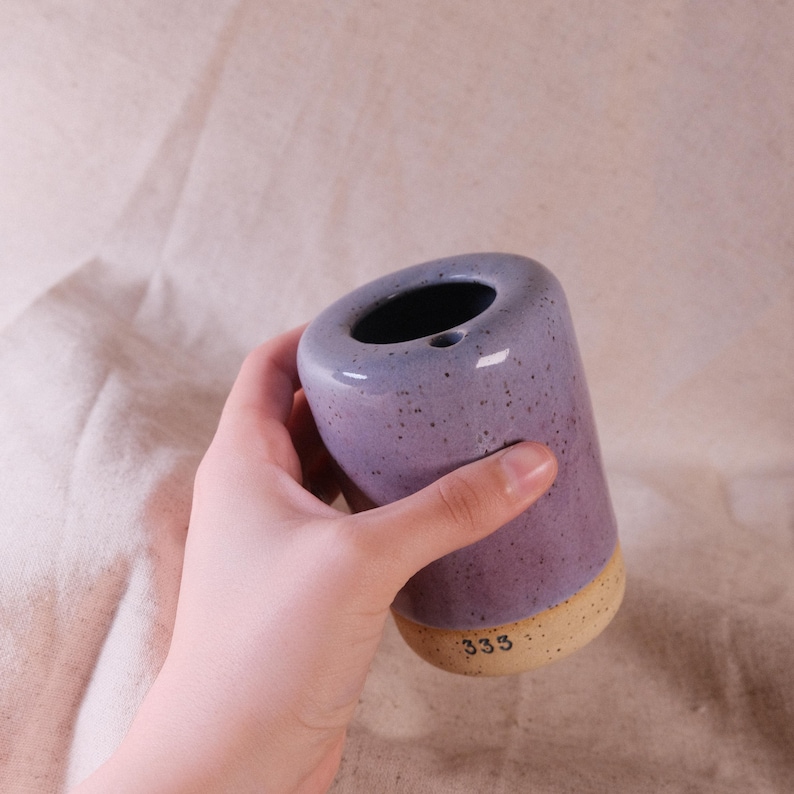 以下が含まれることがあります： 紫とベージュの色合いの円筒形のセラミック花瓶。花瓶は斑点のある仕上がりで、上部に広い開口部があります。底の部分はベージュで、ベースには数字の「333」が印刷されています。花瓶は手で持たれています。