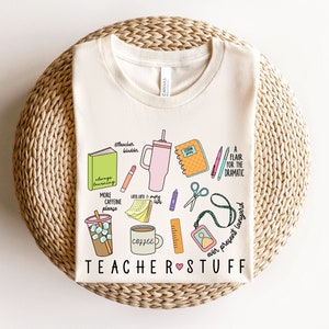 May include: A beige t-shirt with a graphic design of teacher-related items, including a coffee cup, books, pencils, scissors, and a lanyard. The text "TEACHER STUFF" is printed in pink and black.