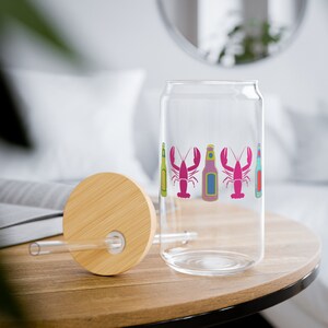 May include: A clear glass can-shaped drinking glass with a bamboo lid and straw. The glass features colourful illustrations of lobsters and beer bottles. The lid and straw are made of bamboo. The glass is on a wooden table.