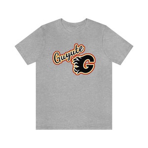 Puede incluir: Una camiseta gris con un gráfico negro y naranja de una "G" estilizada con llamas y el texto "Guyute" encima.