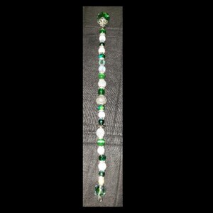 May include: A beaded wand featuring a variety of green and white beads. The wand has a silver-colored metal base and is topped with a green decorative element. The beads vary in size and shape, creating a visually appealing design.