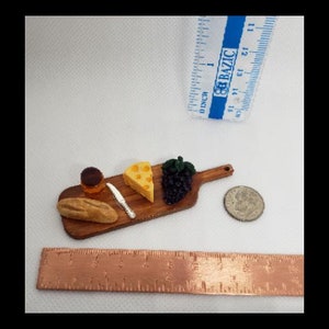 May include: A miniature wooden serving board with a small loaf of bread, a wedge of cheese, a small jar of jam, a bunch of grapes, and a tiny knife.