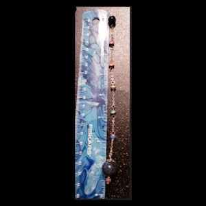 Könnte beinhalten: Eine silberne Kette mit einer blauen Glaskugel am Ende und mehreren kleineren Glasperlen in verschiedenen Farben. Die Kette hängt an einem Lineal.