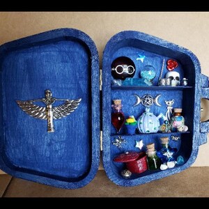 May include: A blue wooden box with a hinged lid, open to reveal a miniature shelf with various decorative items, including a skull, a teapot, a pumpkin, and several small bottles. The box has a silver winged figure on the front.