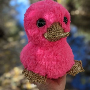 May include: A pink plush toy shaped like a duck with gold glitter accents on the beak and feet. The toy has large black eyes and a fluffy texture.