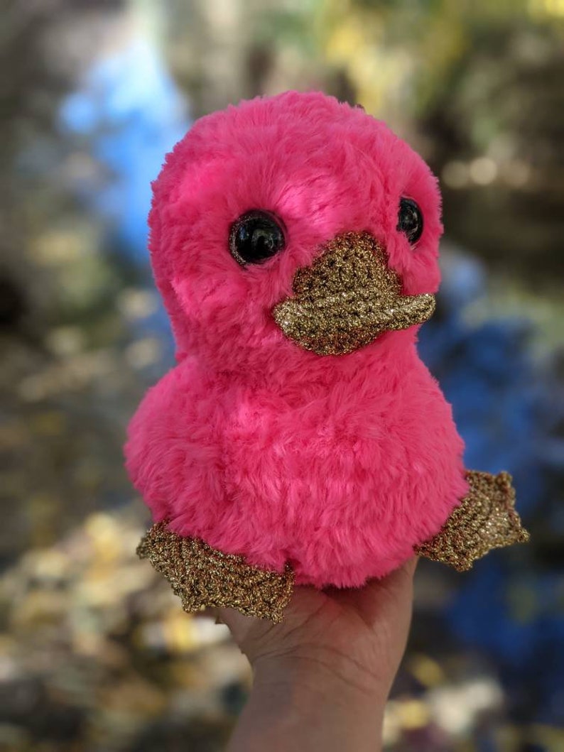 May include: A pink plush toy shaped like a duck with gold glitter accents on the beak and feet. The toy has large black eyes and a fluffy texture.