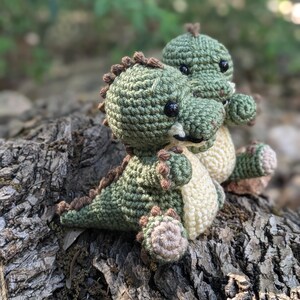 May include: Two crocheted green dinosaur toys with brown accents and black eyes. The dinosaurs are sitting on a tree trunk.