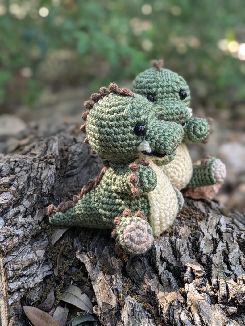 May include: Two crocheted green dinosaur toys with brown accents and black eyes. The dinosaurs are sitting on a tree trunk.