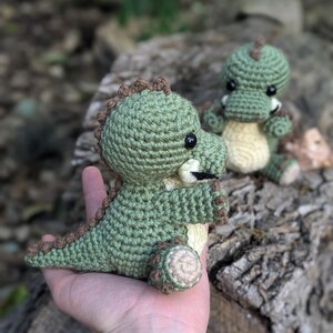 May include: Two crocheted green dinosaur toys with brown accents. The larger dinosaur is being held in a hand, while the smaller dinosaur is sitting on a tree stump.
