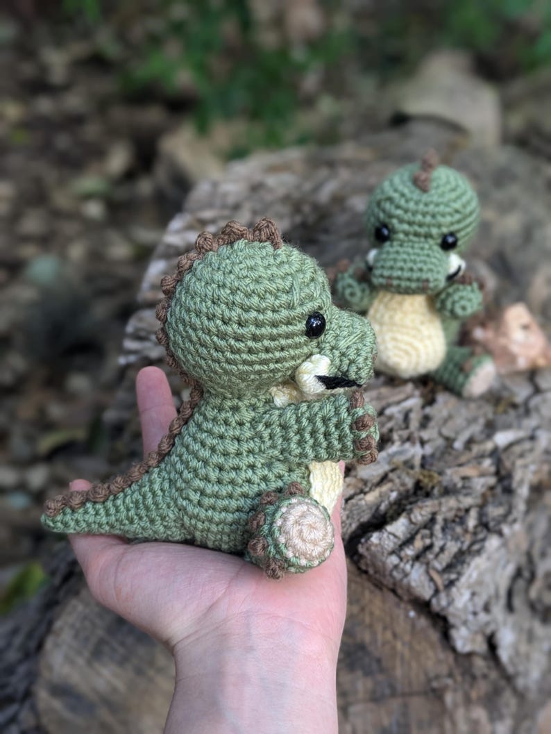 May include: Two crocheted green dinosaur toys with brown accents. The larger dinosaur is being held in a hand, while the smaller dinosaur is sitting on a tree stump.