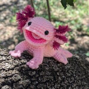 Może przedstawiać: R&oacute;żowa pluszowa zabawka axolotl wykonana na szydełku z czerwonymi, pierzastymi skrzelami. Zabawka siedzi na pniu drzewa.