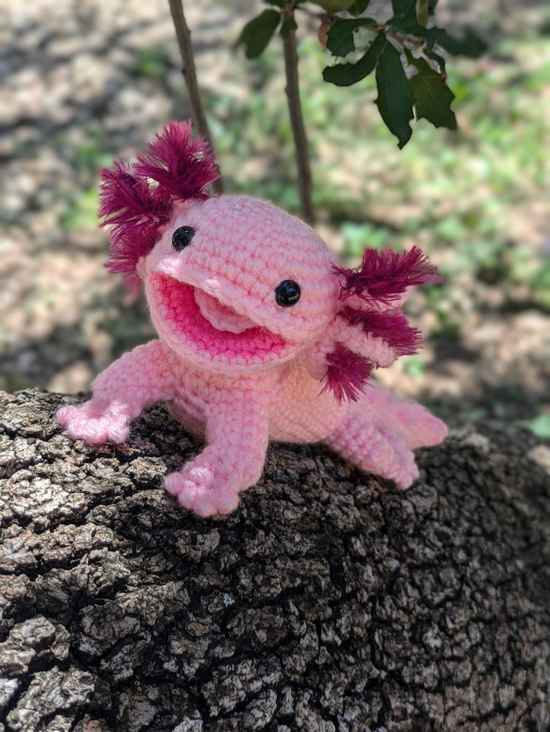 Może przedstawiać: R&oacute;żowa pluszowa zabawka axolotl wykonana na szydełku z czerwonymi, pierzastymi skrzelami. Zabawka siedzi na pniu drzewa.