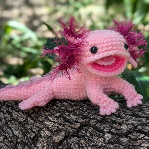 Może przedstawiać: R&oacute;żowa pluszowa zabawka axolotl wykonana na szydełku z r&oacute;żowymi i fioletowymi falbankami. Zabawka ma szeroki, zębaty uśmiech i czarne oczy.