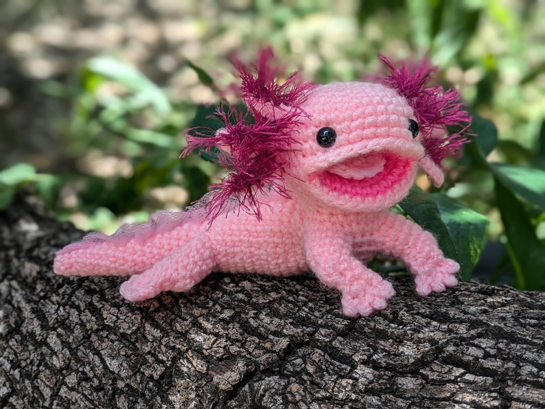Może przedstawiać: R&oacute;żowa pluszowa zabawka axolotl wykonana na szydełku z r&oacute;żowymi i fioletowymi falbankami. Zabawka ma szeroki, zębaty uśmiech i czarne oczy.