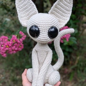May include: A crocheted beige cat with large black eyes. The cat is sitting with its tail curled around its legs.