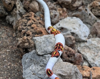 May include: A handmade crochet snake in white with brown, orange, and yellow markings. The snake is posed on a bed of rocks, showcasing its detailed craftsmanship and vibrant colors. The snake's head is black and yellow.